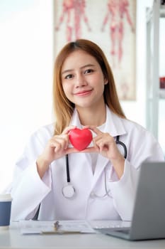 Happy young doctor woman holding red heart shape object, looking at camera with smile. Positive practitioner, cardiologist.