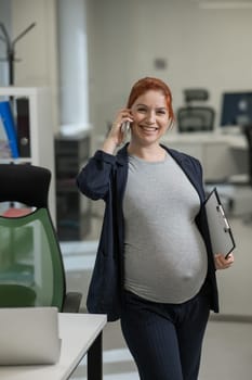 Pregnant woman using mobile phone and holding paper tablet in office. Vertical photo