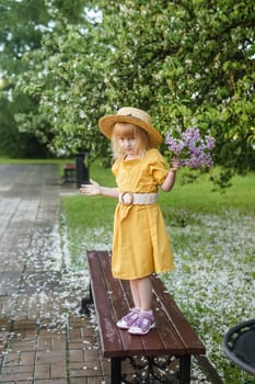 A little girl in a yellow dress and straw hat wearing a bouquet of lilacs. A walk in a spring park, blossoming lilacs