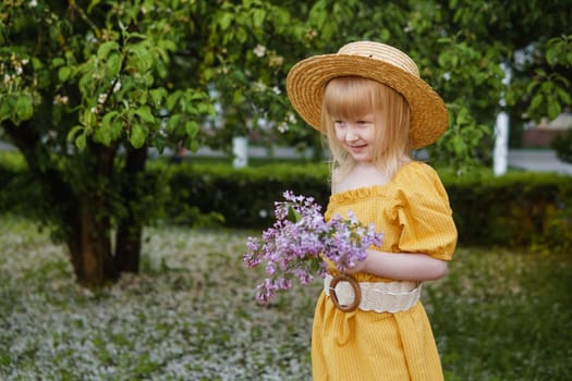 A little girl in a yellow dress and straw hat wearing a bouquet of lilacs. A walk in a spring park, blossoming lilacs