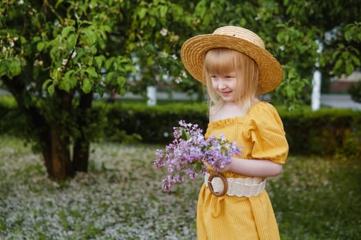 A little girl in a yellow dress and straw hat wearing a bouquet of lilacs. A walk in a spring park, blossoming lilacs