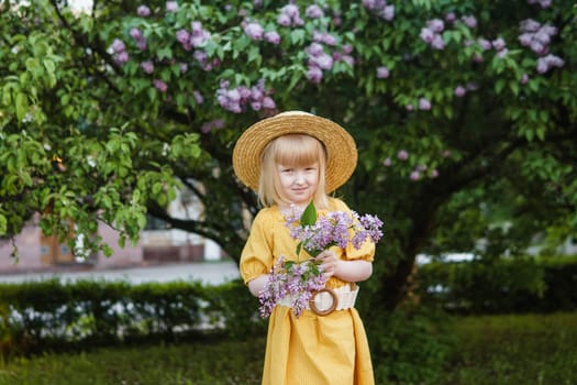 A little girl in a yellow dress and straw hat wearing a bouquet of lilacs. A walk in a spring park, blossoming lilacs