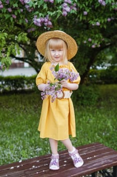 A little girl in a yellow dress and straw hat wearing a bouquet of lilacs. A walk in a spring park, blossoming lilacs