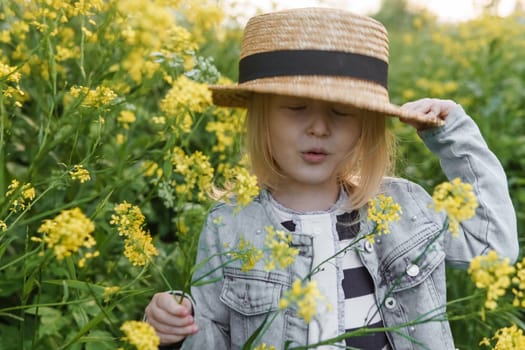 Blonde girl in a field with yellow flowers. A girl in a straw hat is picking flowers in a field. A field with rapeseed