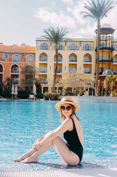 A woman in a black swimsuit, hat and sunglasses is sitting by the pool. The concept of a luxury summer vacation at the hotel.