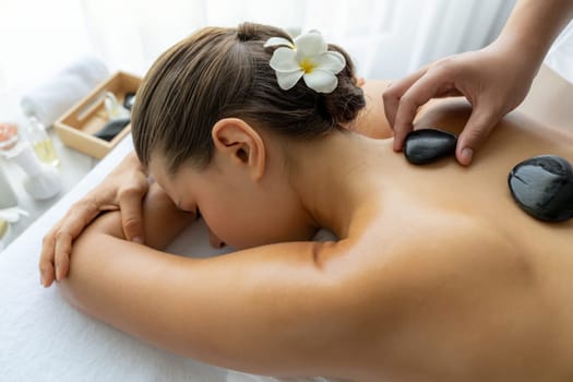 Hot stone massage at spa salon in luxury resort with day light serenity ambient, blissful woman customer enjoying spa basalt stone massage glide over body with soothing warmth. Quiescent