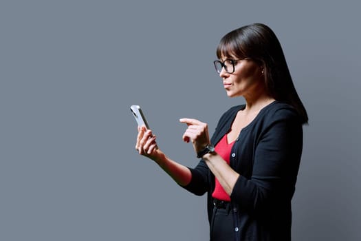 Profile view middle aged serious woman using smartphone on grey background. Mature female looking at phone in hands. Technologies mobile apps applications internet work business leisure communication