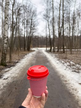 The pink cardboard coffee cup with a red plastic lid in men's hands on the background of a spring park, black jacket, naked forest. High quality photo