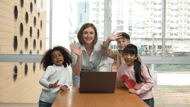 Happy caucasian teacher and multiethnic children waving hands to camera in casual uniform. Smart mentor and diverse student looking at laptop while greeting by moving hands or say good bye. Erudition.