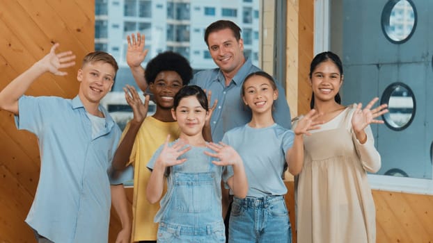 Smart teacher and diverse student standing while waving hand to camera. Skilled instructor with highschool children with mixed races in casual outfit greeting and smiling with cheerful. Edification.