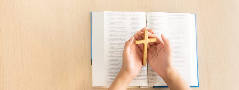 Female god believer holding wooden cross on opened holy bible book at light wooden church table. Top view. Concept of hope, religion, faith, christianity and god blessing. Warm background. Burgeoning.