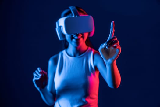 Smart female stand with surrounded by cyberpunk neon light wear VR headset connecting metaverse, futuristic cyberspace community technology. Woman using finger pointing virtual object. Hallucination.