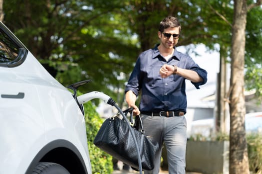 Young man recharge electric car's battery from charging station in outdoor green city park in springtime. Rechargeable EV car for sustainable environmental friendly urban travel lifestyle. Expedient