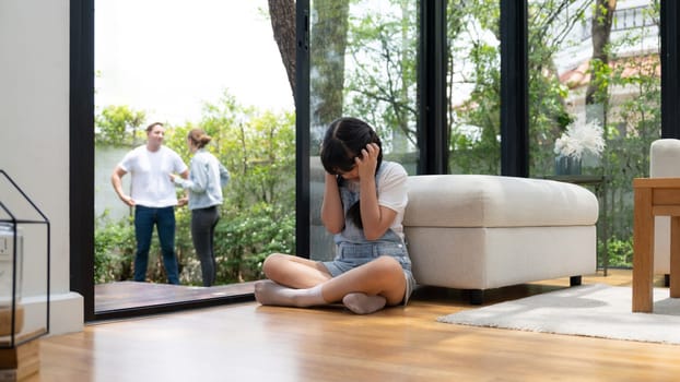 Stressed and unhappy young girl huddle in corner, cover her ears blocking sound of her parent arguing in background. Domestic violence at home and traumatic childhood develop to depression. Synchronos