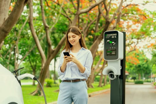 Young woman using smartphone online banking application to pay for electric car battery charging from EV charging station during autumn vacation holiday trip at national park or autumnal forest. Exalt