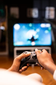 Focus shot on man holding controller in apartment, playing videogames on smart TV display in blurry background. Gamer on sofa using joypad to participate in game on console attached to television set