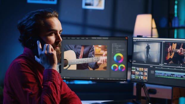 Video editor taking phone call while polishing raw footage, chatting with client about commissioned work. Videographer talks with customer using smartphone, discussing required adjustments