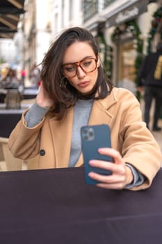 Middle-aged woman using smartphone sitting outdoor cafe. Pretty businesswoman in glasses dressed trench coat holding smartphone use video call to business conversation. Vertical photo