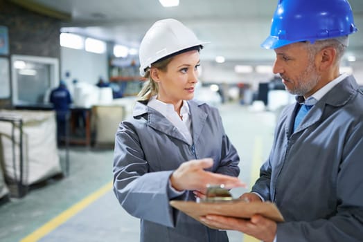 Man, woman and conversation with clipboard in warehouse for quality inspection or safety checks and process monitoring. Team, communication and board for manufacturing information in printing factory.