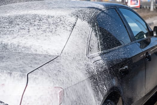 A man washes off the foam while washing the car. A vehicle is engulfed in soapy foam during a car wash, covering the tires, wheels, hood, and entire exterior of the car. Self-service car wash