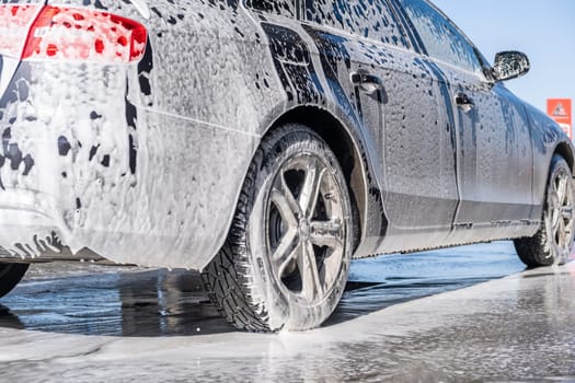 A vehicle is engulfed in soapy foam during a car wash, covering the tires, wheels, hood, and entire exterior of the car. Self-service car wash