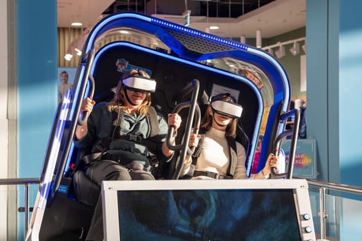 Moscow, Russia, March 14, 2024. Two woman is sitting in an electric blue virtual reality chair, wearing a headset. The machine combines engineering and fun in a display device