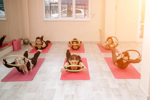 A group of six athletic women doing pilates or yoga on pink mats in front of a window in a beige loft studio interior. Teamwork, good mood and healthy lifestyle concept