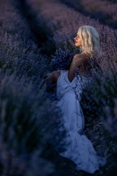 Blonde woman poses in lavender field at sunset. Happy woman in white dress holds lavender bouquet. Aromatherapy concept, lavender oil, photo session in lavender.