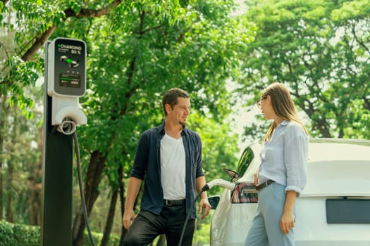 Lovely young couple wearing sun glasses recharging battery for electric car during road trip travel EV car in natural forest or national park. Eco friendly travel during vacation and holiday. Exalt