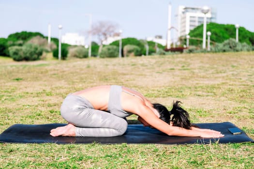 young woman in sportswear kneeling on her yoga mat doing back stretching exercises in the park following the instructions of the app on her mobile phone, active and healthy lifestyle concept