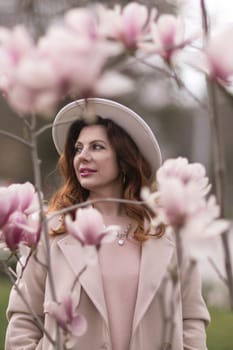 Woman magnolia flowers, surrounded by blossoming trees, hair down, white hat, wearing a light coat. Captured during spring, showcasing natural beauty and seasonal change