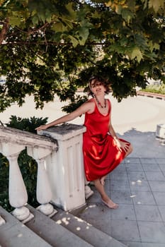 woman in a red silk dress and a bandage on her head smiles against the background of the leaves of a tree. She is leaning on the coop and looking into the camera. Vertical photo