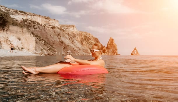 Woman freelancer works on laptop swimming in sea on pink inflatable ring. Pretty lady typing on computer while floating in the sea on inflatable donut at sunset. Freelance, remote work on vacation