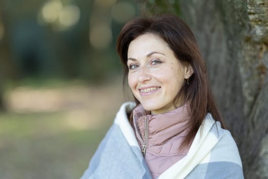 A woman is sitting under a tree and is wearing a pink and white blanket. She is smiling and she is enjoying the outdoors