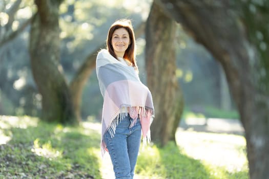A woman is standing in a park, wearing a colorful scarf and jeans. She is smiling and looking towards the camera