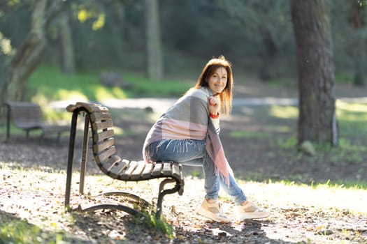 A woman is sitting on a park bench, wearing a scarf and a pink shirt. She is smiling and she is enjoying her time in the park