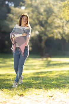 A woman wearing a multicolored scarf is walking in a park. The park is filled with trees and grass, and there is a bench nearby. The woman is enjoying her walk and the peaceful surroundings