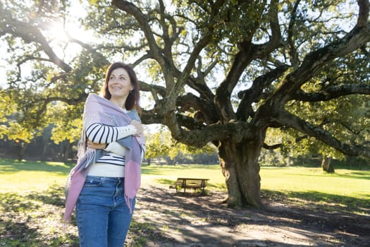 A woman is standing under a tree with a scarf wrapped around her shoulders. The scene is peaceful and serene, with the woman looking up at the tree and smiling