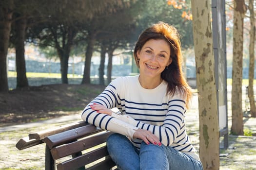 A woman wearing dentistry braces is sitting on a bench in a park. She is smiling and wearing a striped shirt