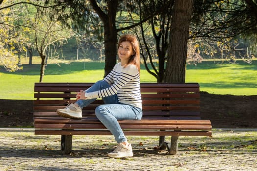 A woman is sitting on a park bench. She is wearing a striped shirt and blue jeans. The bench is wooden and has a few leaves on it. The woman is enjoying her time in the park