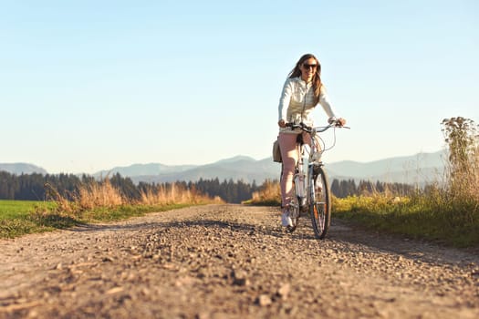 Young long hair woman with sunglasses riding bike on dusty country road, afternoon sun lit country background