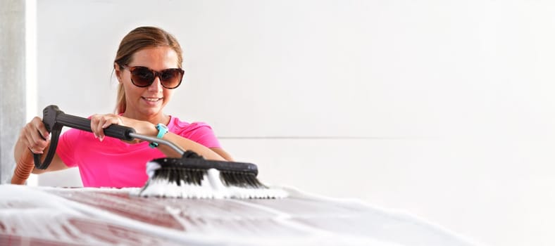 Young woman wearing pink t shirt and sunglasses cleaning her car in self serve carwash, wide banner with space for text right side