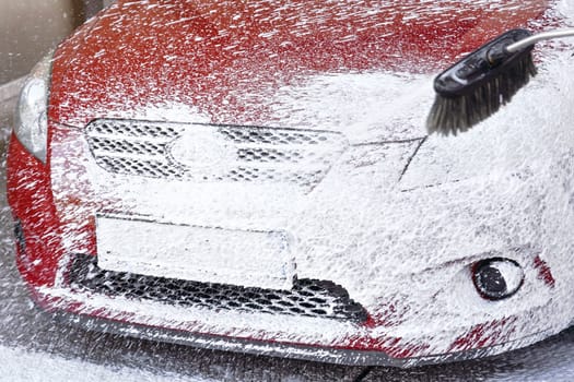 Red car washed in self serve carwash, detail on white soap foam spraying from high pressure hose, covering all front bumper and license plate