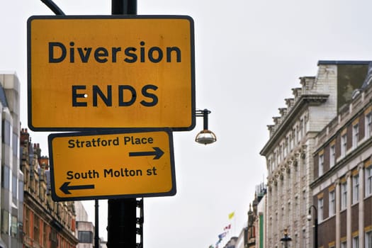 Yellow and black Diversion Ends sign with arrows pointing to Stratford Place and South Molton street, blurred building in background