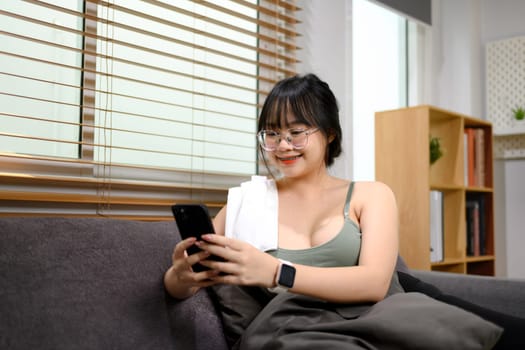 Smiling woman using smartphone resting after fitness training at home.