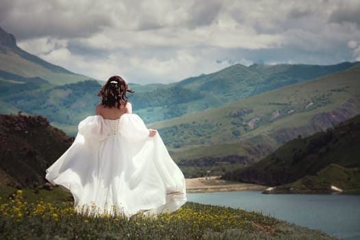 a bride in a white wedding dress walks in the green mountains on the grass, in the background there are mountains and a lake.