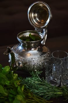 A silver teapot and a bunch of fresh green mint on the foreground. Traditional Moroccan tea as symbol of the hospitality of Moroccan people. Selective focus. Moroccan tea a la menthe
