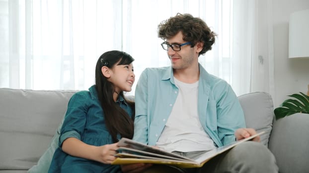 Happy daughter lean on father shoulder while listening story telling. Young asian schoolgirl looking ar dad while reading story book together and sitting at sofa. Family recreation concept. Pedagogy.