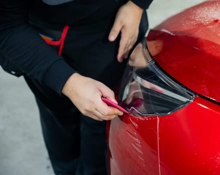 The master applies vinyl film to the headlight of a red car. Closeup view on worker detailer hand smoothing with a scraper protective film
