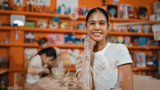 Happy caucasian girl pose at camera while diverse children modeling clay behind. Cute student wearing dirty shirt while looking at camera at workshop in art lesson. Blurring background. Edification.